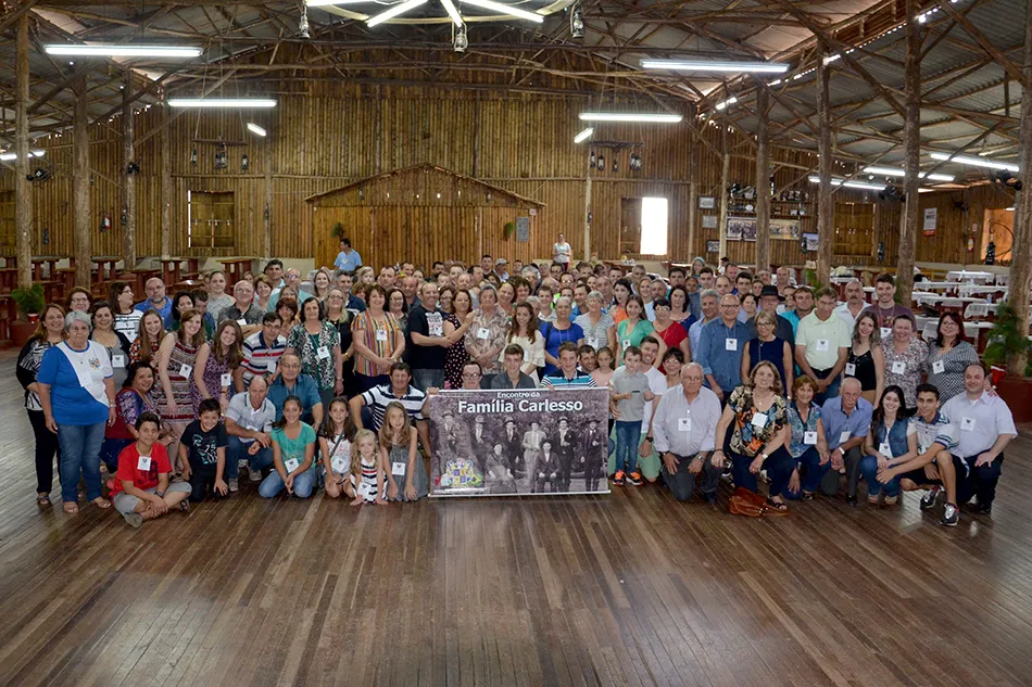 Foto de grupo do 3º Encontro da Família Carlesso, em Chapecó, com cerca de 170 pessoas reunidas num salão de madeira em torno do cartaz da família.